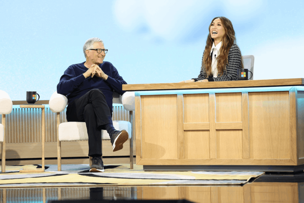 Bill Gates and host Brenda Song sharing a laugh during a Microsoft event interview, sitting on a modern stage with branded mugs and wooden furniture.